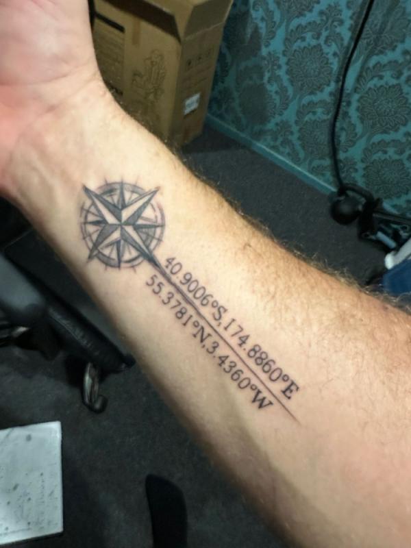 A coordinates tattoo integrated with a compass design on the upper arm, showcasing the precise latitude and longitude coordinates alongside decorative elements.